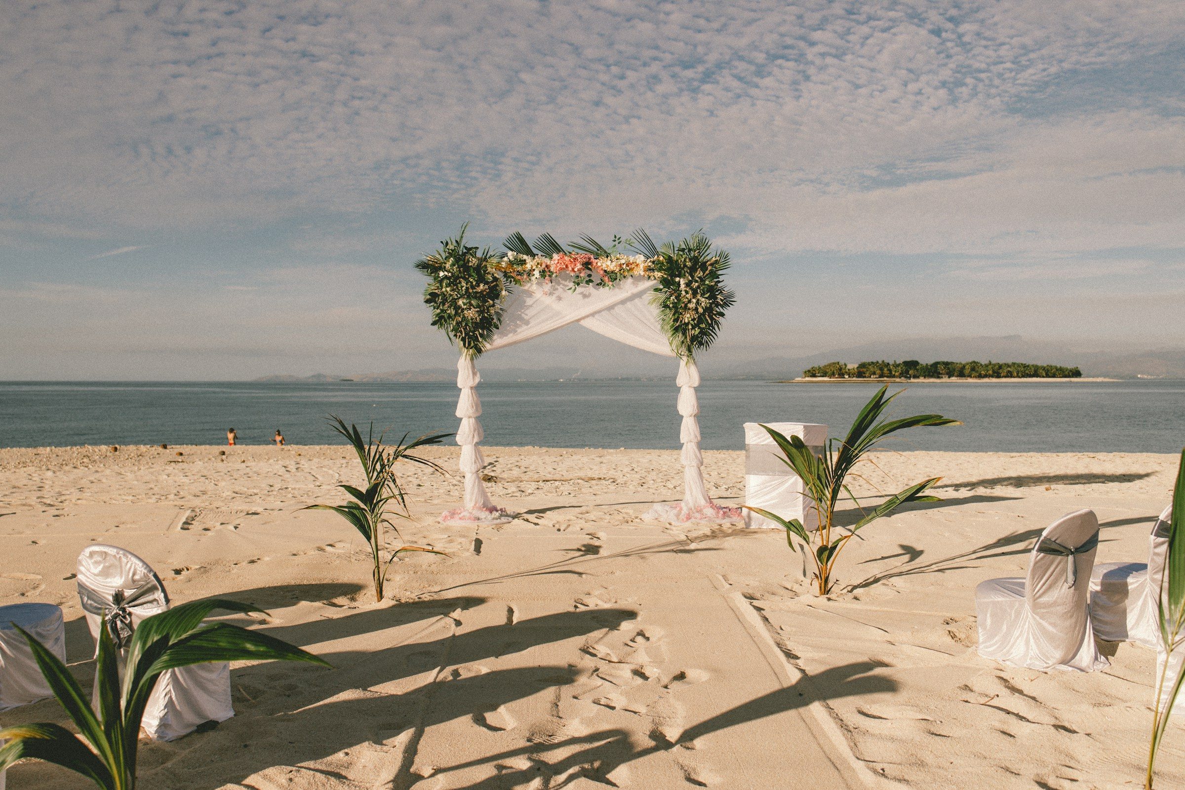 A wedding arch of white flowers and greenery on a sandy beach, an island on the horizon