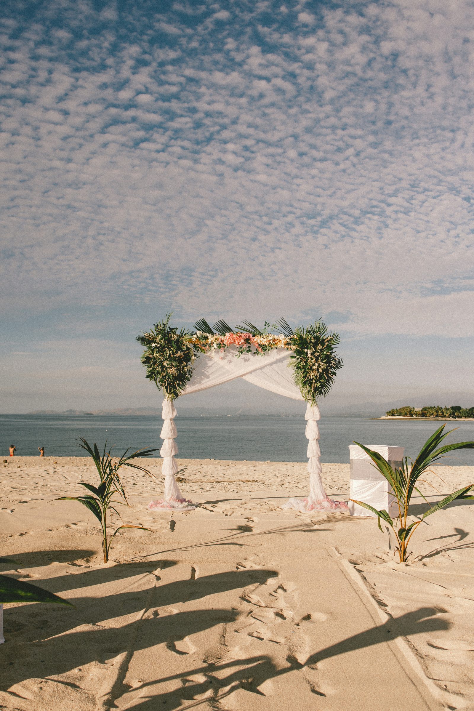 A flower-topped wedding arch and small palms on the beach under a dappled evening sky