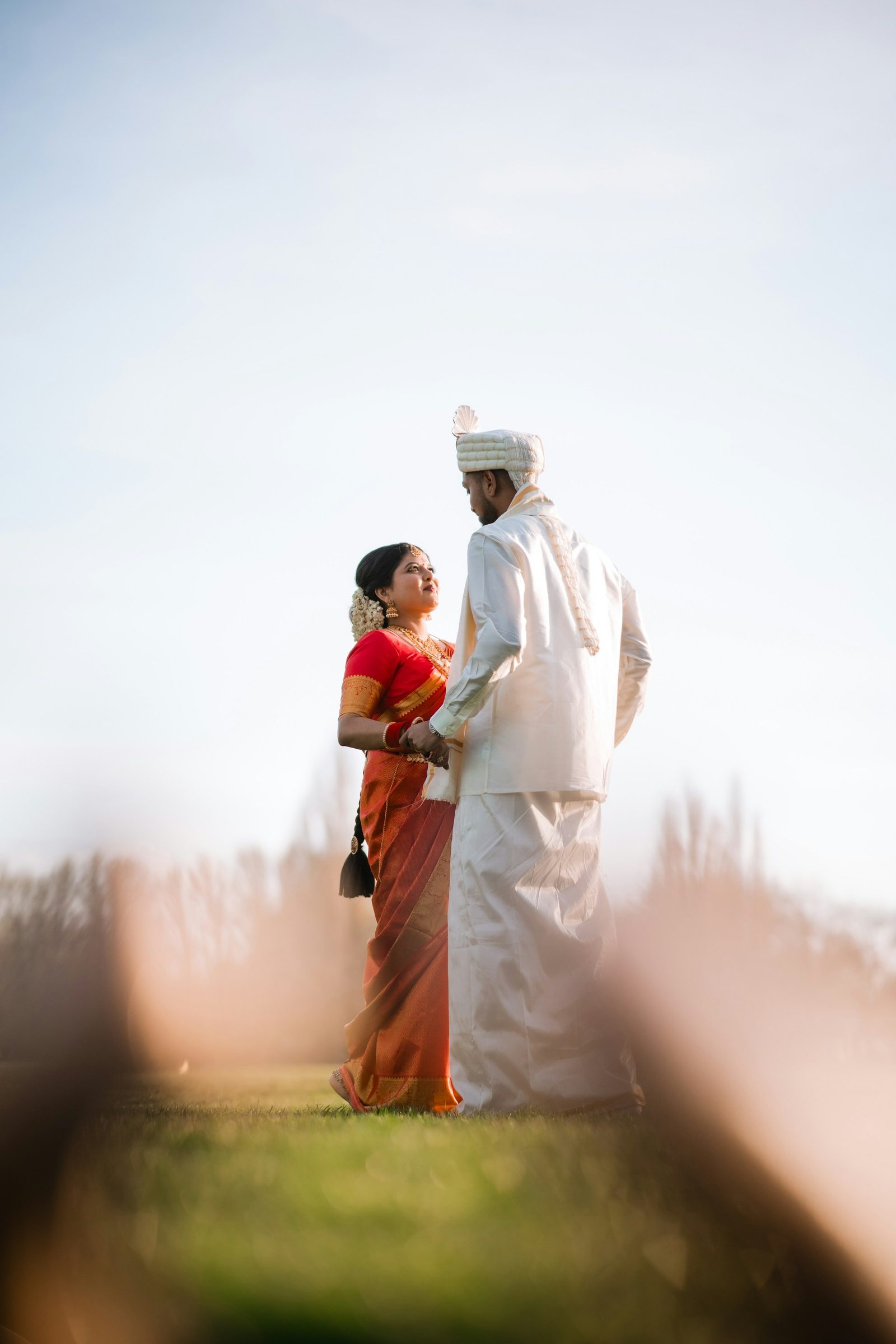 The bride in a red sari and the groom in white stand together in a field at dusk, framed by soft-focus grass