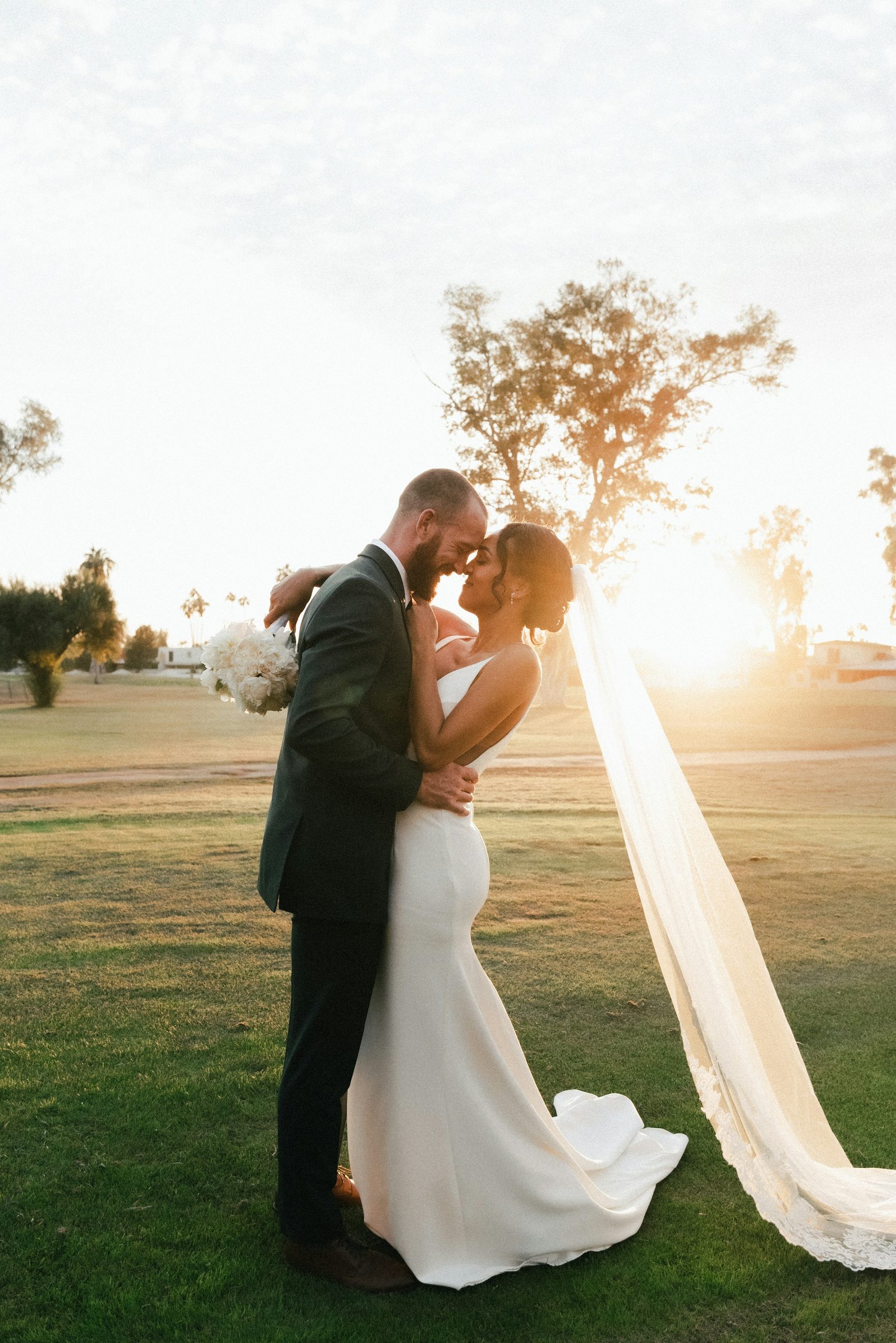 The couple kiss on a lawn as the low sun flares through the bride's long veil