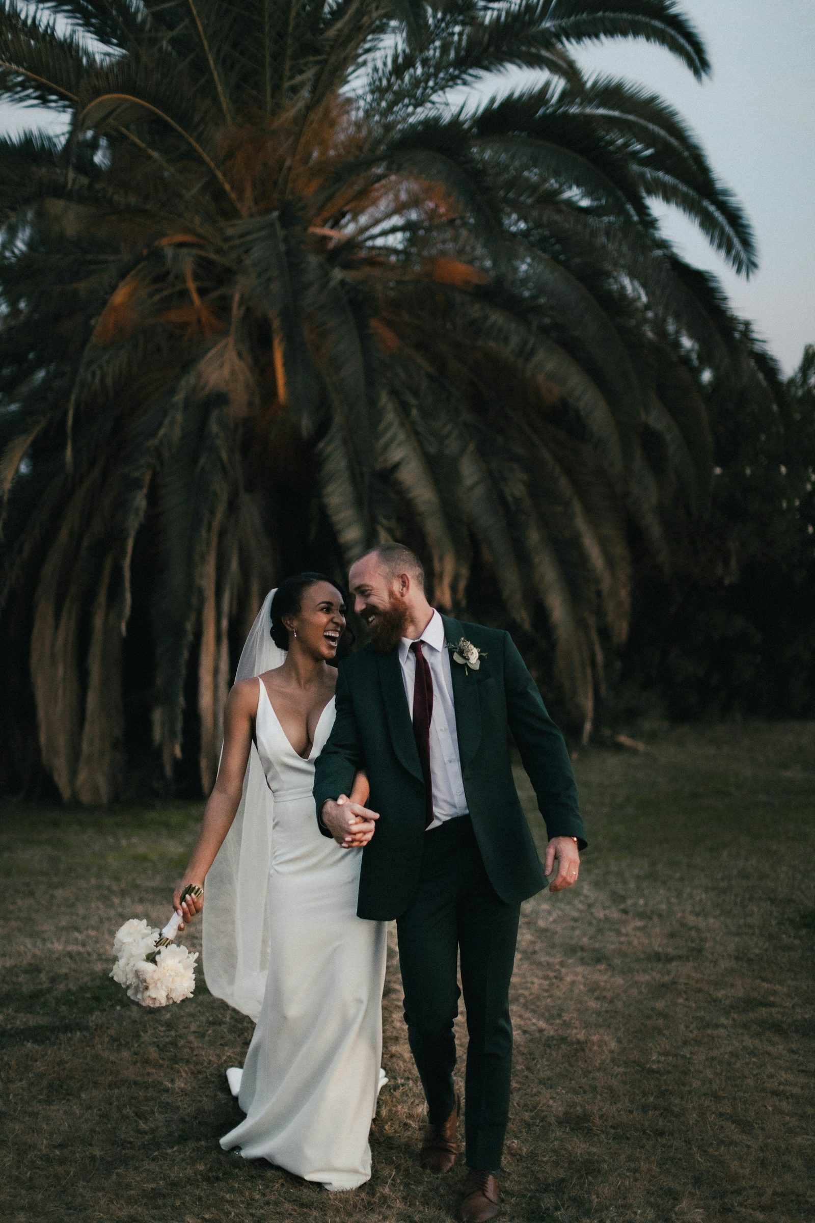 The bride and groom walk arm in arm across the grass beneath a large palm tree